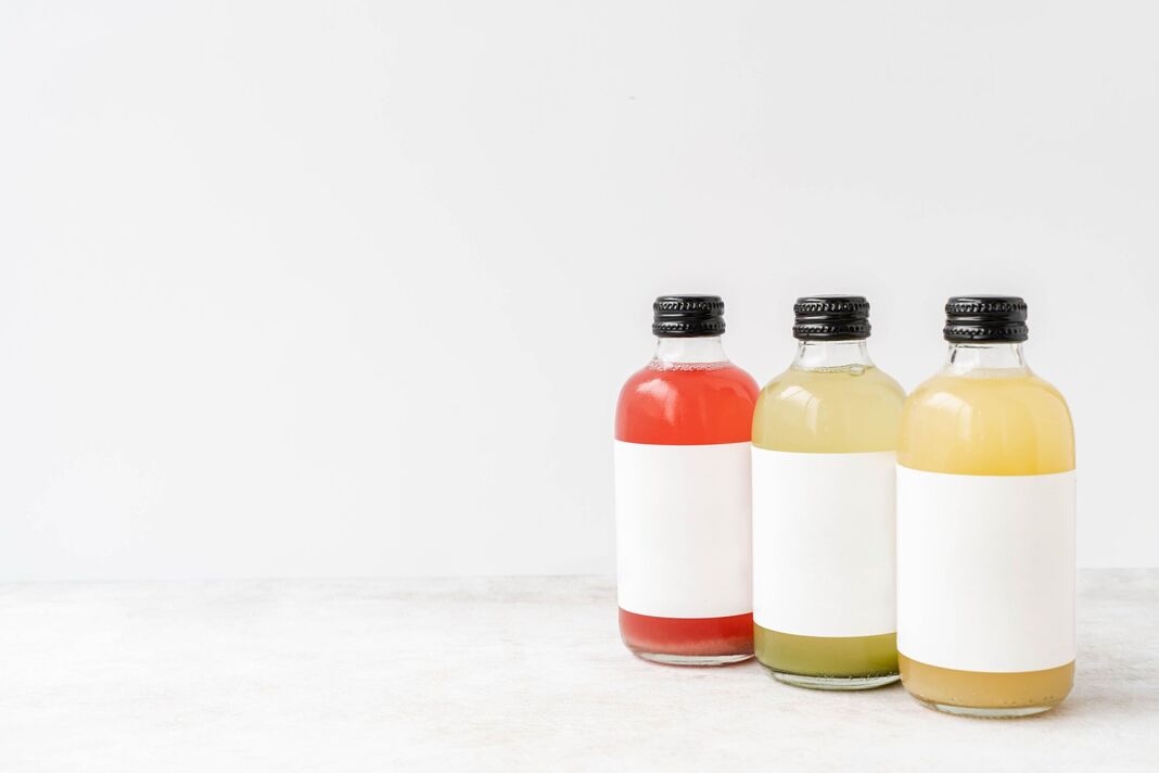 Trois petites bouteilles en verre transparent avec bouchons noirs, remplies de kombucha coloré jaune, rouge et vert