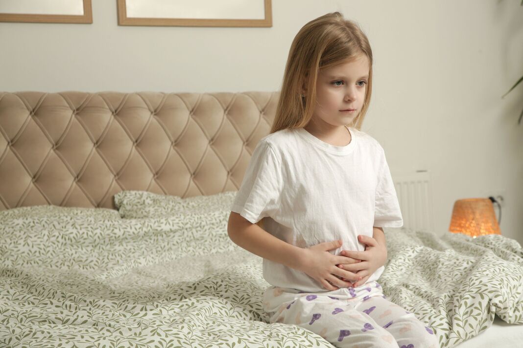 Petite fille assise sur le lit dans la chambre avec des douleurs à l'estomac. Mains sur le ventre.