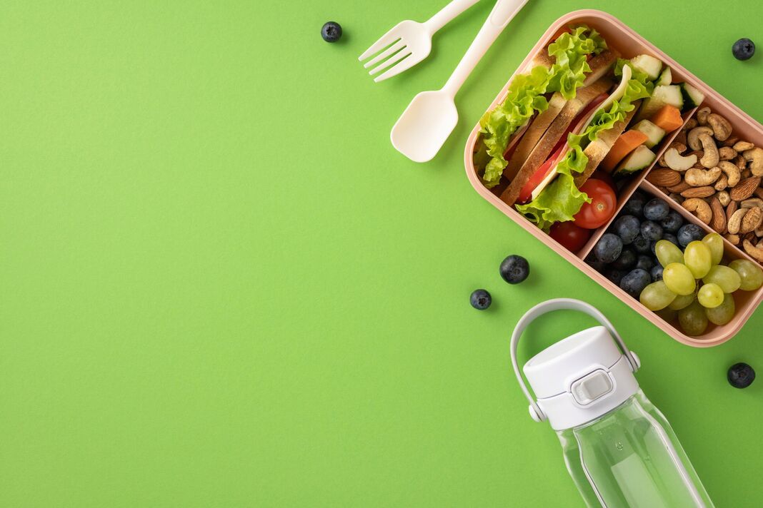 Une vue de haut en bas d'une boîte à lunch saine contenant des fruits frais, des légumes et des noix, accompagnés d'une bouteille d'eau sur un fond vert vibrant. Idée