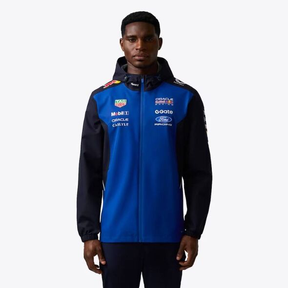 Un individu vêtu d'une veste de sport bleue et noire avec une capuche, arborant divers logos et textes de sponsors, pose pour une photo